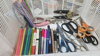 Photo showing assortment of colorful pens, markers, multiple scissors, hole punchers, and dog nail clippers inside a white plastic basket.