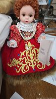 Close-up of Queen Elizabeth I doll in red velvet dress with gold embroidery holding an identification card.