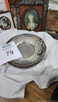 Photo of the silver saucer with floral decoration and beaded edge, displayed on white cloth with framed art behind it.