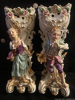 Front view of figurative porcelain vases depicting young woman and young man in period costumes with raised floral and scroll detailing and gold highlights.