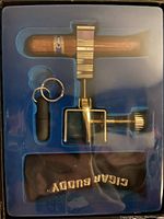 Photo showing the Cigar Buddy set in original packaging containing a brass cigar cutter with clamp holding a cigar, a punch with keychain, and a black branded pouch.