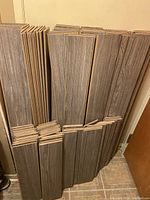Stack of greyish hardwood flooring planks leaning against a wall showing wood grain texture and thickness.