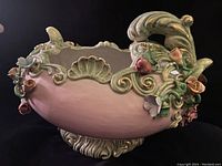 Side view of scalloped oval ceramic pedestal bowl with ornate floral and scroll decorations in pastel colors (green, pink, blue).