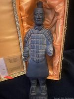Black terracotta warrior figure statue with detailed armor, standing on a rectangular base, inside satin-lined decorative box with patterned trim.