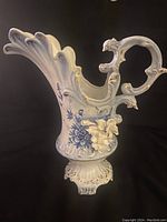 Ceramic pitcher front view showing ornate handle, blue floral design, and flared spout.