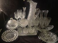 Full lot of various vintage clear glassware including a tall textured pitcher, covered dish, multiple glasses, and serving bowls and dishes arranged on a dark surface.