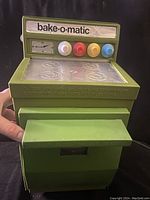 Front view of the green Bake-O-Matic toy oven showing control panel with four colored knobs and branding.