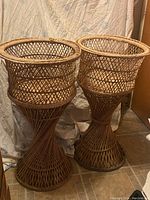 Photo showing two vintage wicker plant stands next to each other against a backdrop. Both stands have a woven lattice wicker design with hourglass shape and round top basket.