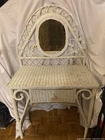 Front view of white wicker vanity showing the oval mirror, tabletop, single drawer, and ornate wicker scrollwork decor.