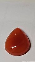 Photo of pear-shaped polished mandarin orange carnelian cabochon gemstone lying flat on surface, showing smooth convex shape and glossy finish.