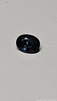 Photograph of the natural blue sapphire gemstone placed on a white surface showing oval cut and deep blue color.