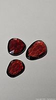 Three fancy cut natural cherry red garnet gemstones displayed on a light surface, showing their deep red color and faceted cuts in heart and pear shapes.