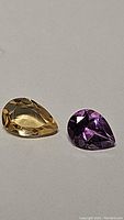 Photo showing pear shaped golden citrine and deep purple amethyst gemstones side by side on a neutral background.