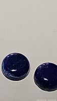 Close-up photo of two deep blue round cabochon lapis lazuli gems on neutral background showing smooth glossy surface.