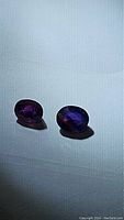 Two oval-cut multicoloured sapphires showing rich purple and blue to pink and orange hues with visible inclusions under direct light on a white background.