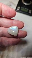 Close-up photo of a natural rainbow moonstone held between fingers, showing iridescent blue flash and slight imperfections on the side.