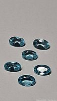 Six oval cut paraiba blue apatite gemstones arranged on gray surface, showing top and angled views.