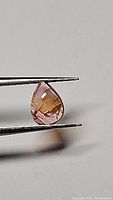 Pear-shaped pink orange tourmaline cabochon held in tweezers, showing front condition and color detail of the gemstone.