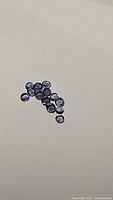 Group of approximately thirteen cleaned and polished round cabochon purple iolite gemstones, each about 3mm diameter, placed on a white background.