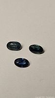 Three oval faceted sapphires: two teal green stones and one blue stone on a plain background, showing color and cut details.
