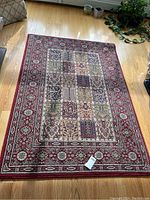 Full view of the Valby Ruta area rug from an elevated angle showing the detailed patchwork pattern and the red floral border laid on a wooden floor with some room decor visible.