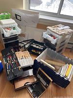 Overview of multiple plastic bins, brush cases, wooden easel and palette on floor