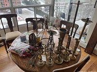 Wood table with multiple brass candlesticks of different sizes and styles, metal angel, candle snuffers, bag of colored taper candles, and jar candles in glass containers. Chairs and balcony visible in background.