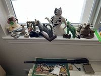 Shelf with various decorative figurines: cat in dress, striped cat on wooden base, black cat, green frog, and small animal figurines.