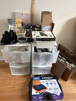 Full view of lot items stacked, showing storage drawers with items on top including Kabooti cushion box, binoculars, headphones, and boxes of lightbulbs and small household items.