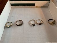 Five assorted ladies rings laid out in a cardboard box, showing variety of designs and size