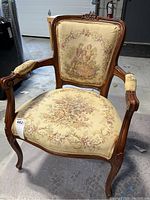 Front view of the antique occasional arm chair showing carved wood and detailed upholstery with couple and floral tapestry.
