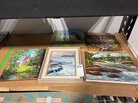 Lot of five original paintings on canvas and board displayed on cardboard on shelf. Includes framed and unframed pieces.