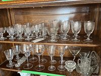 Photo showing two shelves of assorted clear crystal and glass stemware including wine glasses and goblets of various styles and sizes, and decorative bowls on a wooden cabinet shelf.