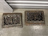 Two antique cast iron vent covers with ornamental frame and central decorative grille, shown side by side on the floor.