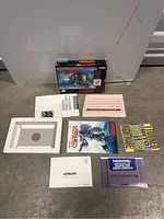Layout of the Cybernator SNES game cartridge, instruction booklet, box, and various paper inserts on floor