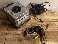 Silver Nintendo GameCube console with power adapter and composite AV cable on wooden surface.