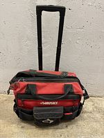 Front view of red and black Husky rolling tool bag with extended handle, displaying front pockets and logo.