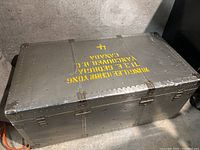 Photo showing the large metal chest with riveted construction and yellow stenciled text on lid.