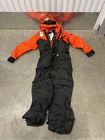 Front view of orange and black anti-exposure coverall laid flat, showing hood, sleeves, torso and legs