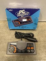Photo of gray NEC PC Engine TurboPad controller laid flat in front of its original blue retail box showing front view of controller and box artwork.