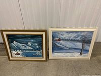 Two framed winter landscape paintings placed on floor against wall.