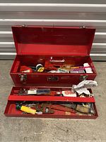 Red metal tool chest open, showing the removable upper tray with various tools including pipe wrench, hammer, pliers, tape measure, utility knife, and other items.