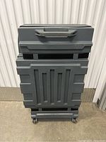 Front view of gray plastic trade show case with handle on top and wheels at the bottom, showing reinforced ridged design.