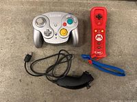 Photo showing silver Wavebird GameCube controller, red Wii remote with blue wrist strap, and black Wii nunchuk controller on concrete floor.