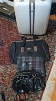 Overview of four bags: large white suitcase, black duffel bag, Burberry-style plaid tote, black leather handbag placed on patterned carpet.