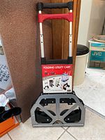 Front view of folding utility cart with red and black extendable handle and folded base, showing product tag with weight capacity and usage examples.