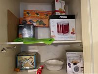 Shelf view showing boxed frozen yogurt maker, pasta maker box, dolma wrapper apparatus box, Braun citromatic juicer box, green kitchen tool, kitchen scale and white colander.