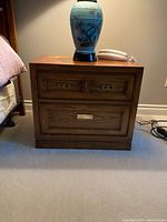 Front view of one wooden nightstand showing the two smaller drawers on top and one larger drawer below with brass-colored handles, medium brown wood grain finish, placed on carpeted floor next to bed and table lamp.