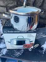 Shiny stainless steel 6.5 quart Dutch oven with lid placed on top of its box outside on snow.