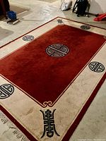 Photo showing the entire large area rug laid flat on a floor, red central field, beige border with black circular and geometric medallions, fringed edges visible.